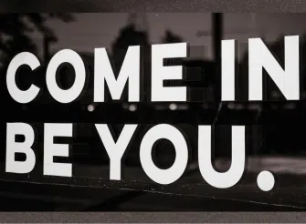 Scritta bianca “COME IN. BE YOU.” su sfondo scuro, simbolo del concetto di autenticità e dell’invito a essere se stessi nella comunicazione.